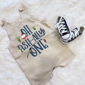 May include: A beige baby romper with the text "Oh Fish-Ally One" in blue and fishing-themed illustrations. Accompanied by navy blue and white canvas trainers. The romper has button fastenings on the shoulders.