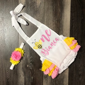 May include: White onesie with yellow and pink ruffles, a bee graphic, and the text "One Bianca". A matching yellow and pink headband with a pink flower is included.