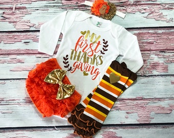 May include: A white long-sleeved onesie with the text "My first thanks giving" in gold glitter lettering. The onesie is paired with an orange ruffled diaper cover with a gold bow, and brown and orange striped leg warmers with a gold heart.