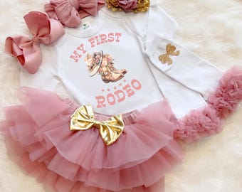 May include: A white baby bodysuit with the text "MY FIRST RODEO" and a graphic of a cowboy hat and boots. The outfit includes a pink tutu skirt with a gold bow, a matching headband, and a gold floral headband.