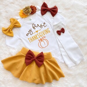 Könnte beinhalten: Ein weißer Langarmbody mit dem Text my first THANKSGIVING und einem Kürbis-Motiv, ein senfgelber Rock mit einer dunkelroten Schleife, eine senfgelbe Haarschleife, eine dunkelrote Haarschleife, ein Blumenstirnband und weiße Stulpen mit einer kleinen Schleife.