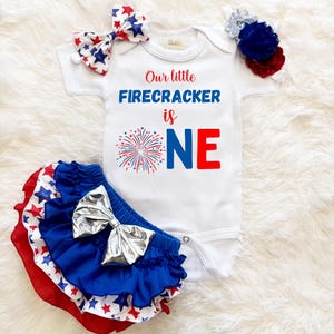 May include: A baby's patriotic outfit set. It includes a white onesie with the text "Our little FIRECRACKER is ONE", a red, white, and blue tutu, and matching hair accessories. The outfit has a festive design with star and firework graphics.