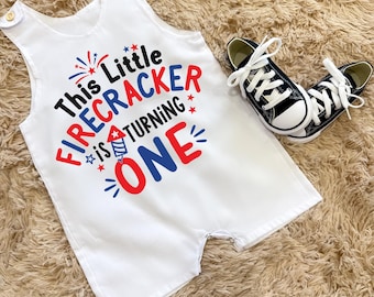 May include: White romper with the text "This Little Firecracker is Turning ONE" in red, blue, and black. Includes a pair of black and white canvas sneakers. The romper has button closures on the shoulders.