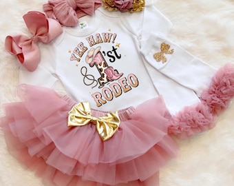May include: A baby girl's first birthday outfit set. The set includes a white long-sleeved bodysuit with a "Yee Haw! 1st Rodeo" graphic, a pink tutu skirt with a gold bow, and matching accessories like bows and a headband.