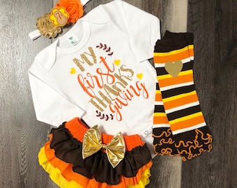 May include: A baby outfit set for Thanksgiving. It includes a white bodysuit with the text "My first Thanksgiving" in gold glitter, a brown, orange, and yellow ruffled skirt with a gold bow, and striped orange, brown, and white leg warmers with a gold heart.