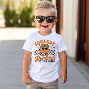 Può includere: Un bambino indossa una maglietta bianca con la scritta "Coolest Pumpkin in the Patch". La maglietta presenta una zucca dei cartoni animati con occhiali da sole e un cappello. Il design utilizza colori arancione, marrone e nero.