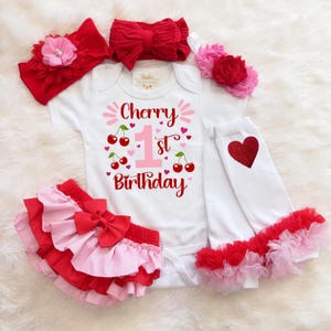 Może przedstawiać: Białe body dla dziecka z napisem "Cherry 1st Birthday" i grafiką wiśni. Zestaw zawiera czerwoną i różową spódniczkę z falbanami, białe legginsy z czerwonym sercem oraz czerwone i różowe opaski na głowę z kokardkami i kwiatami.