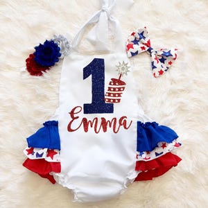 May include: White romper with red, white, and blue detailing, featuring a large number 1 and the name "Emma" in glitter. Includes a matching headband and bow, ideal for a first birthday celebration.