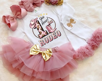 May include: A baby girl's rodeo-themed outfit. The set includes a white onesie with a "Yee Haw! 1st Rodeo" graphic, a pink tutu skirt with a gold bow, and matching accessories like a headband and bows. The outfit is in shades of pink and gold.