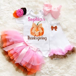 Könnte beinhalten: Ein weißer Kurzarm-Body mit dem Text Sophia's 1st Thanksgiving und einem Kürbis-Motiv, ein mehrlagiger Tüll-Tutu in Pink, Orange und Braun, weiße Stulpen mit einer glitzernden Schleife, eine rosa Haarschleife und ein Blumen-Stirnband.