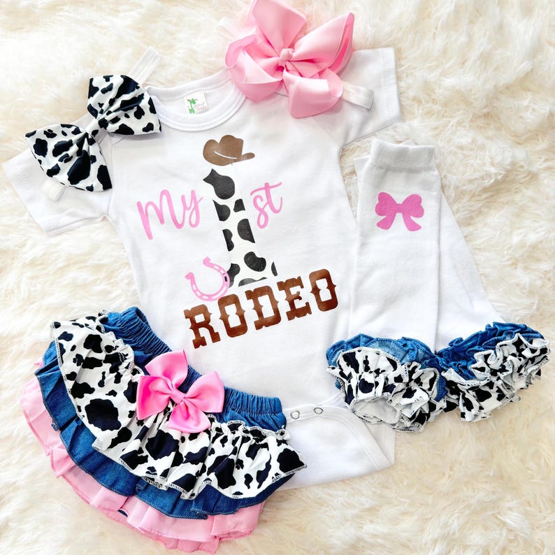 May include: A white onesie with the text "My 1st Rodeo" in pink and brown lettering. The onesie features a brown cowboy hat and a cow print boot. The outfit includes a pink bow headband, a cow print and denim ruffle skirt, and pink and white leg warmers with a pink bow.
