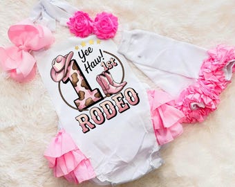 May include: White baby bodysuit with pink ruffle trim and a matching pink bow. The bodysuit features a cowboy hat and boot graphic with the text "Yee Haw! 1st Rodeo". Includes a white headband with pink flowers.