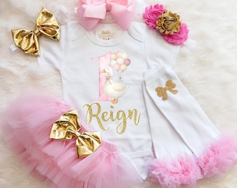 May include: A white baby outfit set with a pink tutu, leggings, and bows. The set includes a onesie with a duck graphic and the number 1. Gold glitter accents are used throughout the set, including the name "Reign".