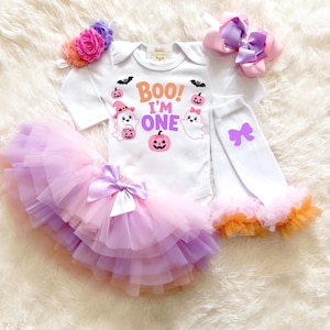 May include: A white long-sleeve bodysuit with the text "BOO! I'M ONE" and ghost and pumpkin graphics, paired with a layered tulle tutu in shades of pink and purple, a floral headband, a large hair bow, and white leg warmers with a purple bow.