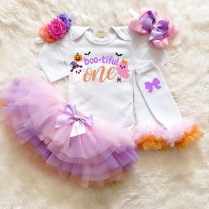 May include: A white long-sleeve bodysuit with the text "boo-tiful one" featuring a pumpkin, ghosts, and bats. Includes a layered tulle tutu in shades of pink, purple, and orange with a satin bow, a floral headband, a hair bow, and leg warmers.