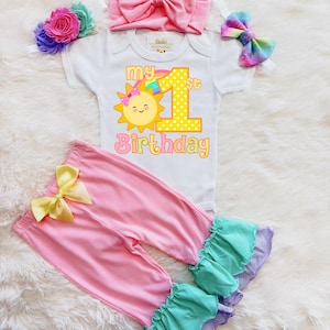 May include: A baby's first birthday outfit set. The set includes a white onesie with a sun graphic and the text "My 1st Birthday", pink pants with a yellow bow, a pink headband, and other accessories.