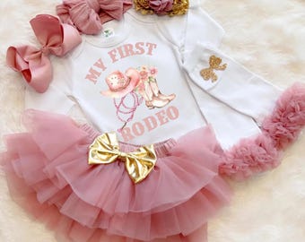 May include: A baby girl's rodeo-themed outfit set. The set includes a white onesie with a pink graphic, a pink tutu skirt with a gold bow, and matching pink hair accessories. The onesie says "My First Rodeo".
