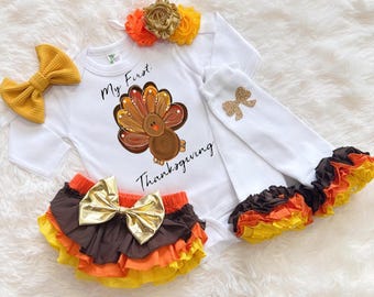 May include: A white baby bodysuit with a turkey graphic and the text "My First Thanksgiving". The set includes a yellow bow, ruffled bloomers, and leg warmers with bows and ruffles in fall colors. A floral headband completes the outfit.