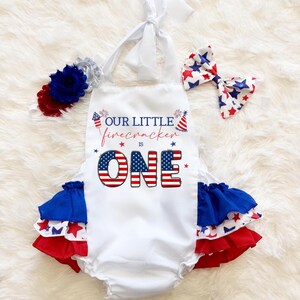 May include: A white baby romper with the text "OUR LITTLE firecracker IS ONE" in red, white, and blue. The romper has ruffled trim in red, white, and blue. A matching headband and bow complete the patriotic outfit.
