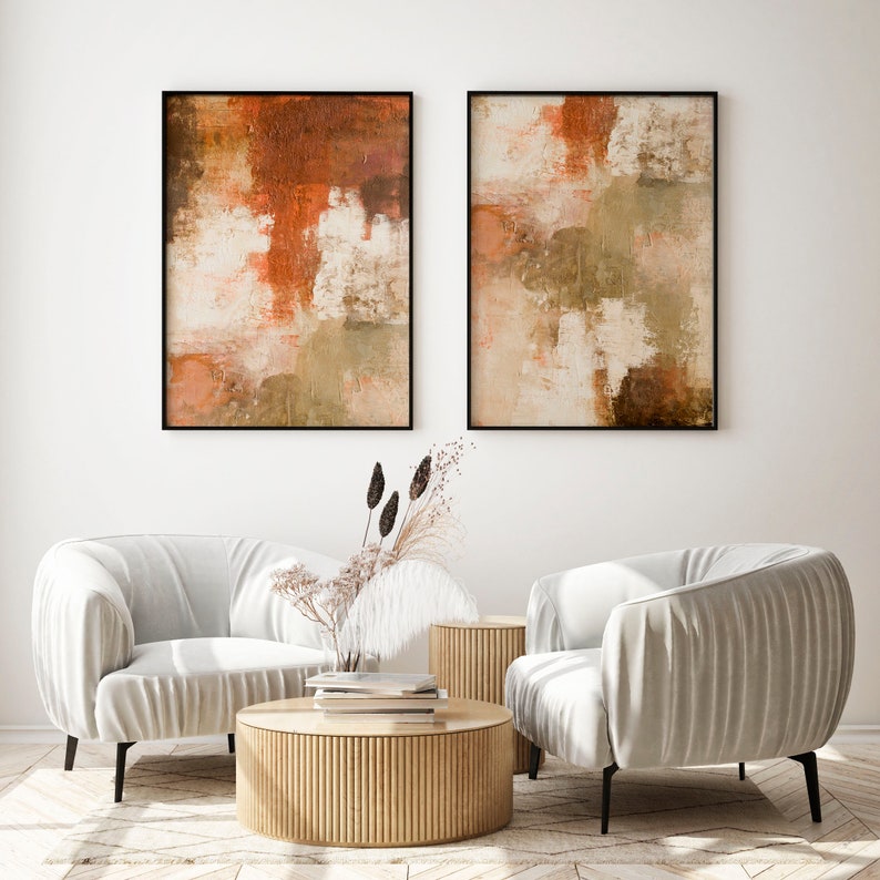 May include: Two abstract paintings in black frames with muted orange, brown, and beige colors hang above two white armchairs with a wooden coffee table in front of them.