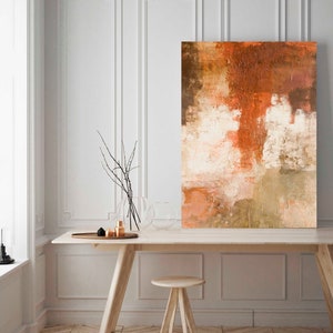 May include: Abstract painting with shades of brown, orange, and white. The painting is hanging on a white wall above a wooden table with a vase of branches and a few decorative objects.