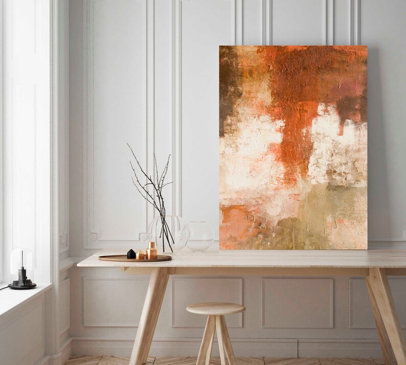 May include: Abstract painting with shades of brown, orange, and white. The painting is hanging on a white wall above a wooden table with a vase of branches and a few decorative objects.