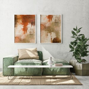 May include: Two abstract paintings in white frames, one with orange and brown hues, the other with green and brown hues, hanging on a white wall above a green sofa with a beige pillow and a white coffee table with a vase and a bowl.