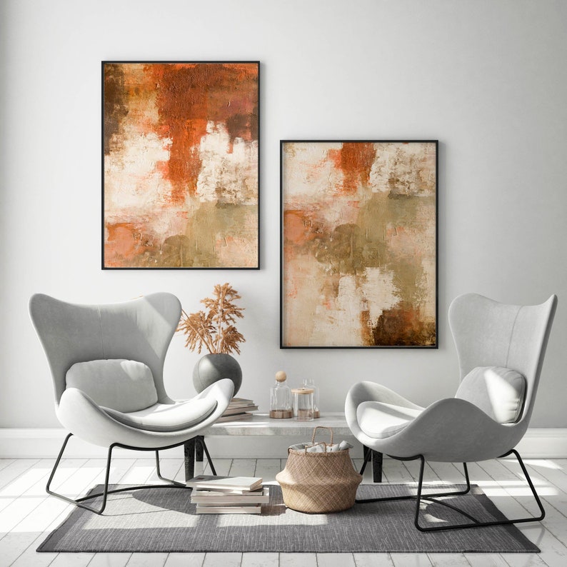 May include: Two abstract paintings in warm tones of brown, orange, and white, hanging on a white wall above two gray wingback chairs. The chairs are facing each other and are positioned in front of a coffee table with a woven basket and decorative objects.