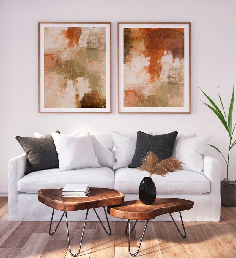 May include: Two abstract paintings with brown, orange, and white colors hang above a white couch with pillows and two wooden coffee tables with black metal legs.