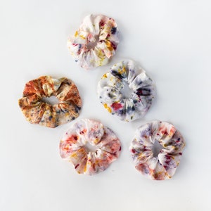 Puede incluir: Cinco scrunchies de tela coloridos con un patr&oacute;n de tie-dye en tonos de amarillo, naranja, rojo, morado y azul. Los scrunchies est&aacute;n dispuestos en un patr&oacute;n triangular sobre un fondo blanco.