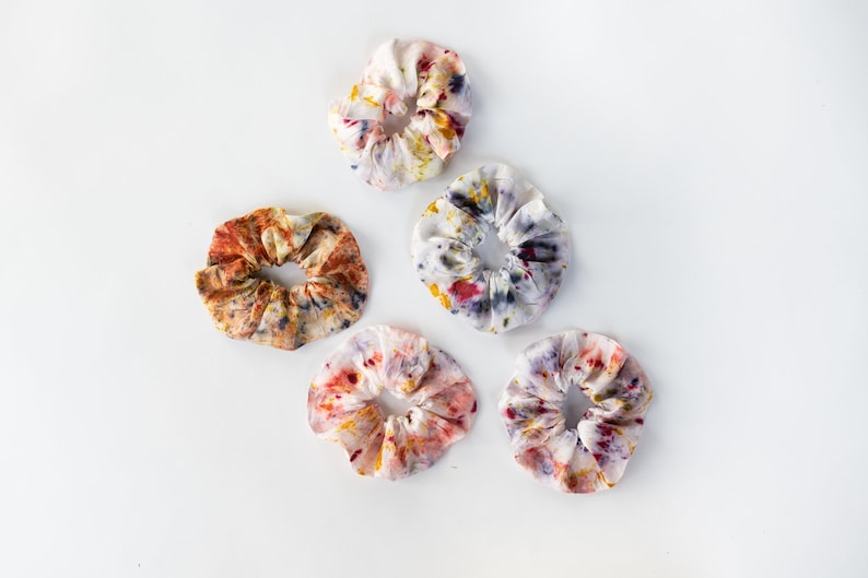 Puede incluir: Cinco scrunchies de tela coloridos con un patr&oacute;n de tie-dye en tonos de amarillo, naranja, rojo, morado y azul. Los scrunchies est&aacute;n dispuestos en un patr&oacute;n triangular sobre un fondo blanco.