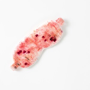 May include: A white sleep mask with a pink and red watercolor design. The mask has a textured surface and is decorated with small red dots.