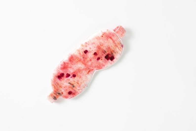 May include: A white sleep mask with a pink and red watercolor design. The mask has a textured surface and is decorated with small red dots.