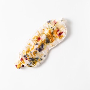 May include: A white silk sleep mask with a colorful floral pattern in shades of yellow, orange, red, and purple.
