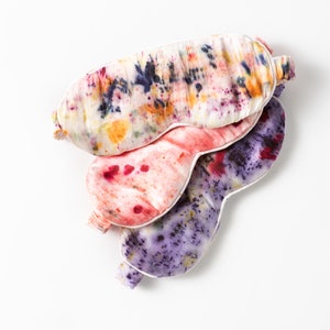 May include: Three colorful silk sleep masks with tie-dye patterns in shades of purple, pink, and yellow. The masks are stacked on top of each other.