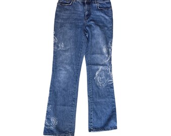 May include: Light blue denim jeans with a bootcut leg. The jeans feature a white paisley design on the front and back. The jeans have a classic five-pocket design and a button and zipper closure.