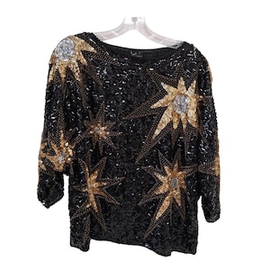 May include: Black sequined top with gold and silver starburst designs. The long-sleeved top is embellished with sequins and beads, creating a shimmering effect. The top is hanging on a clear plastic hanger.