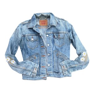 May include: Light blue denim jacket with a classic collar, button closure, and multiple pockets. The jacket features white daisy flower patches on the sleeves. A small red tag is visible on the chest pocket.