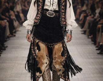 May include: A model wearing a Western-inspired outfit on a runway. The ensemble includes a black vest with floral embroidery, a white blouse, a black skirt with cowhide accents, and snakeskin boots. The outfit is accessorized with a belt and jewelry.