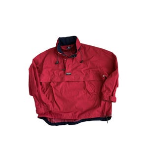 May include: A red pullover windbreaker jacket featuring a navy blue inner collar, a partial front zipper, and a horizontal flap pocket across the chest. The jacket includes elasticized cuffs, a drawstring hem, and a small rectangular logo patch.
