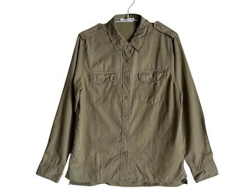 Puede incluir: Camisa verde oliva con botones, mangas largas, dos bolsillos en el pecho y charreteras. La camisa está colgada de una percha de metal sobre un fondo blanco. La camisa tiene un diseño clásico y utilitario.