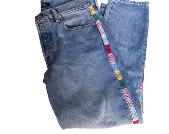 May include: Light blue denim jeans with a colorful patterned stripe down the side. The jeans have a classic five-pocket design and a faded wash. The decorative stripe features various colors and geometric patterns.