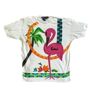 May include: White t-shirt featuring a vibrant flamingo design. The flamingo is pink with black accents, set against a backdrop of a palm tree and a colorful geometric border. The shirt has a crew neck and short sleeves.