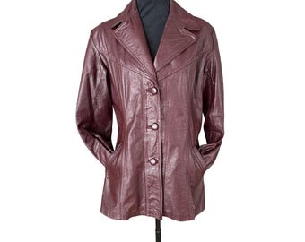 Può includere: Giacca in pelle bordeaux con colletto a revers e chiusura a tre bottoni. La giacca ha due tasche anteriori ed è esposta su un manichino nero. La giacca è di colore pieno e sembra essere vintage.