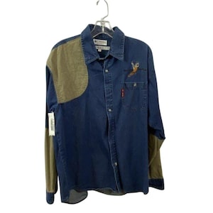 May include: A long-sleeved denim shirt with olive green shoulder and elbow patches. The shirt has an embroidered bird design on the front pocket and a small red tag. The shirt is hanging on a hanger.