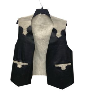 May include: A black leather vest with a light grey suede-like yoke and pocket accents. The sleeveless vest hangs on a black hanger. The vest has a classic western design.
