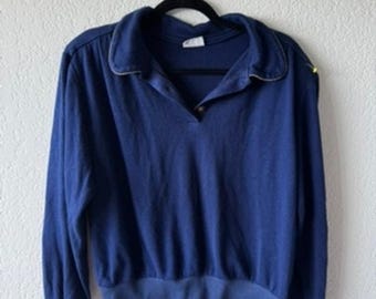 Könnte beinhalten: Ein marineblaues Langarmshirt mit Kragen und V-Ausschnitt. Das Shirt hat einen goldenen Besatz am Kragen und einen Knopfverschluss. Die Manschetten und der Saum sind gerippt. Das Shirt hängt an einem schwarzen Kleiderbügel.
