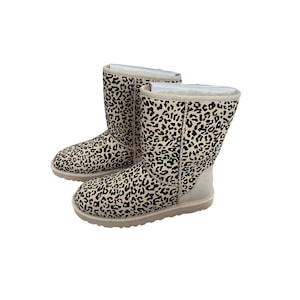Puede incluir: Un par de botas con estampado de leopardo beige y negro. Las botas son de media caña con punta redonda y suela de color claro. Las botas tienen un aspecto suave y texturizado, sobre un fondo blanco.