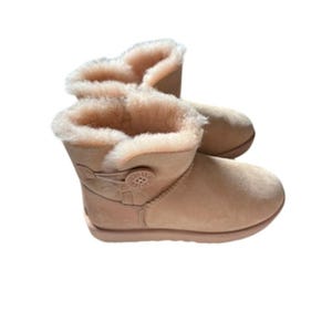 Könnte beinhalten: Ein Paar hellbraune Wildleder-Ankle Boots mit Knopfdetail an der Seite. Die Stiefel haben ein weiches, flauschiges, hellbraunes Innenfutter und eine runde Spitze. Die Stiefel sind für kaltes Wetter konzipiert.