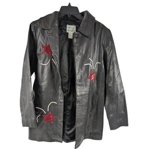 May include: Black leather jacket with red star and white accent embroidery. The jacket has a button-up front, a collar, and a black interior lining. The jacket is hanging on a black hanger.